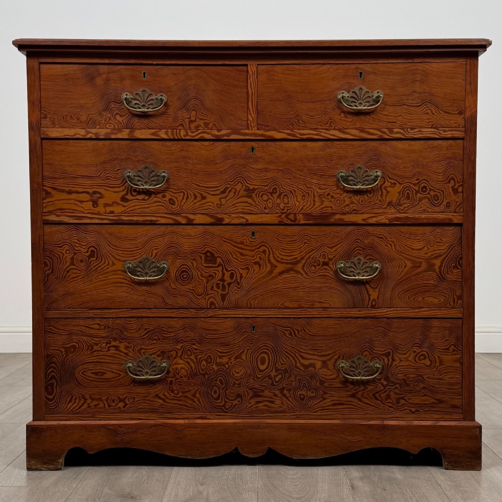 A Victorian nineteenth-century pitch pine chest of drawers featuring a prominent figured wood grain, brass drop handles, a two-over-three drawer configuration, and a shaped apron base.