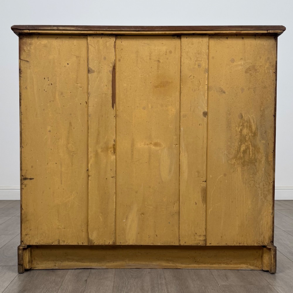 A Victorian nineteenth-century pitch pine chest of drawers featuring a prominent figured wood grain, brass drop handles, a two-over-three drawer configuration, and a shaped apron base. View V