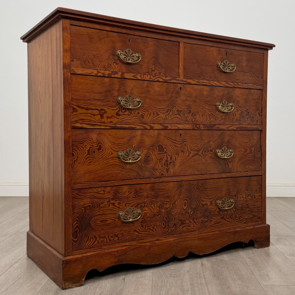 A Victorian nineteenth-century pitch pine chest of drawers featuring a prominent figured wood grain, brass drop handles, a two-over-three drawer configuration, and a shaped apron base. View VIII