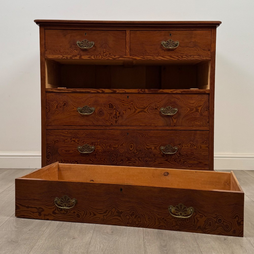 A Victorian nineteenth-century pitch pine chest of drawers featuring a prominent figured wood grain, brass drop handles, a two-over-three drawer configuration, and a shaped apron base. View X