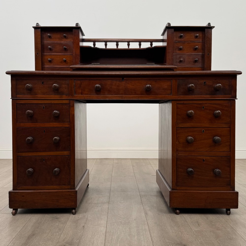 A 19th-century Victorian mahogany Dickens desk featuring a twin-pedestal design, raised superstructure with drawers and spindle gallery, slant writing surface, turned timber knobs, and plinth bases.