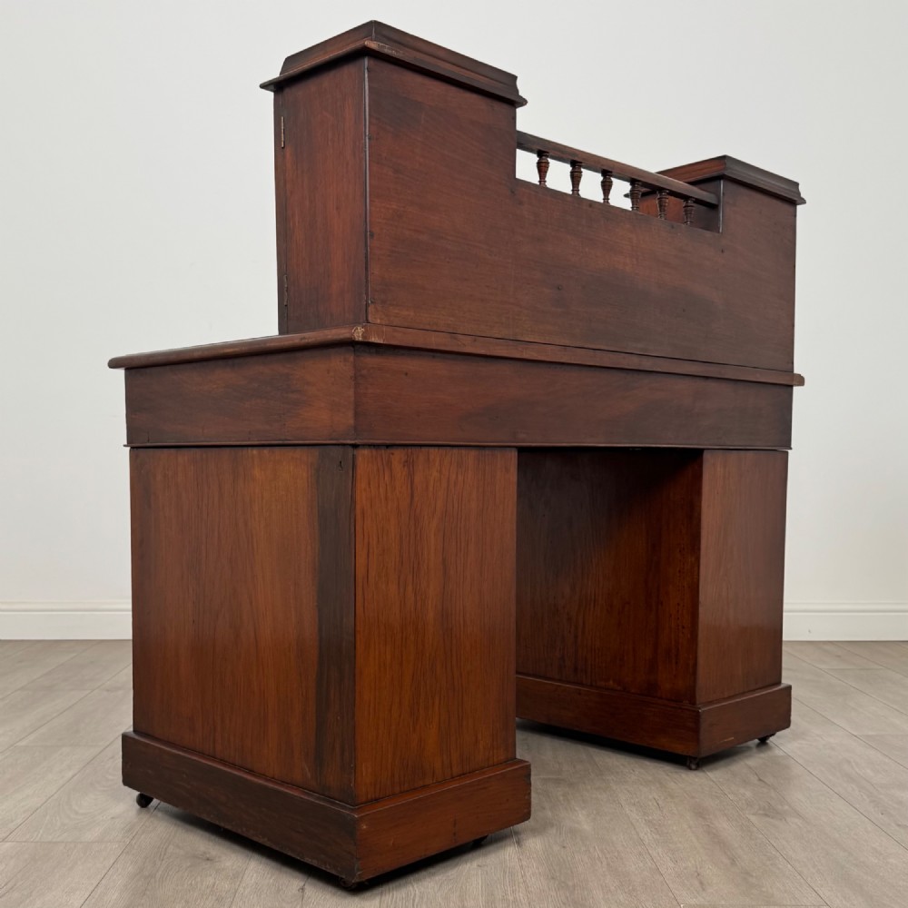 A 19th-century Victorian mahogany Dickens desk featuring a twin-pedestal design, raised superstructure with drawers and spindle gallery, slant writing surface, turned timber knobs, and plinth bases. View V