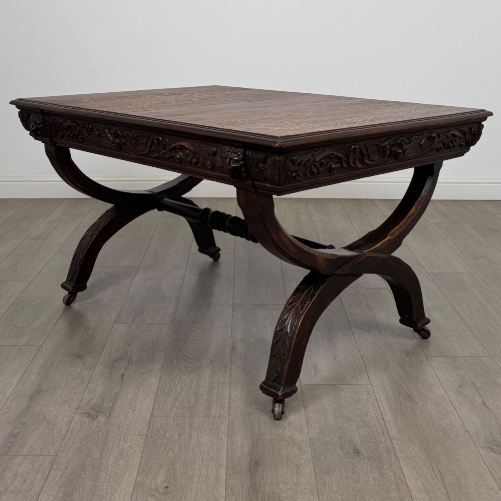 A 19th-century dark carved oak centre table featuring an X-frame curule base, carved frieze, turned stretcher, and metal castors.