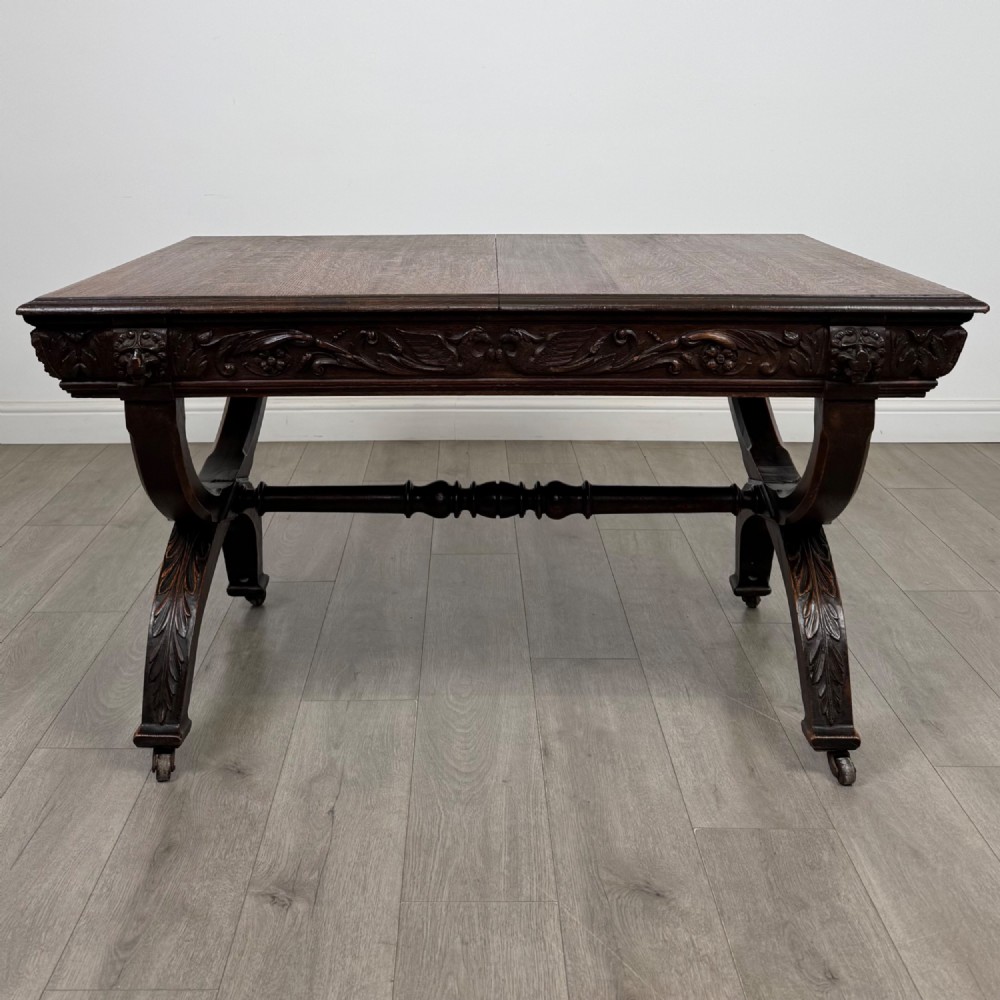 A 19th-century dark carved oak centre table featuring an X-frame curule base, carved frieze, turned stretcher, and metal castors. View II