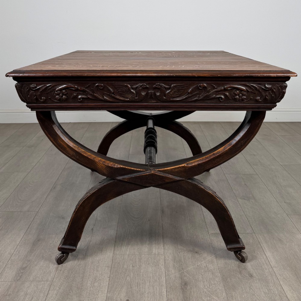 A 19th-century dark carved oak centre table featuring an X-frame curule base, carved frieze, turned stretcher, and metal castors. View III