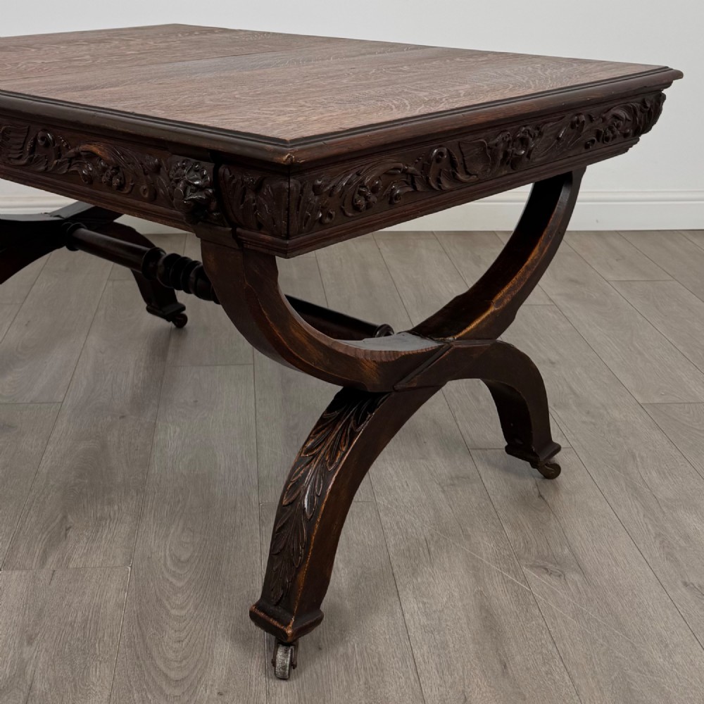 A 19th-century dark carved oak centre table featuring an X-frame curule base, carved frieze, turned stretcher, and metal castors. View IV