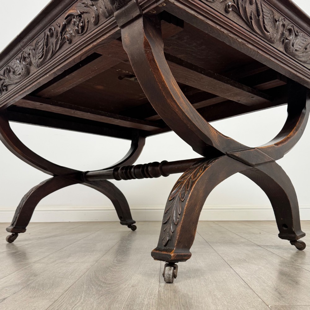 A 19th-century dark carved oak centre table featuring an X-frame curule base, carved frieze, turned stretcher, and metal castors. View V