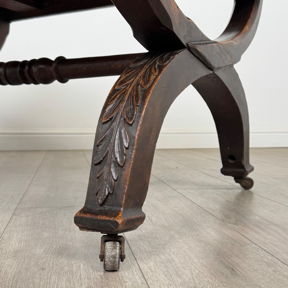 A 19th-century dark carved oak centre table featuring an X-frame curule base, carved frieze, turned stretcher, and metal castors. View VII