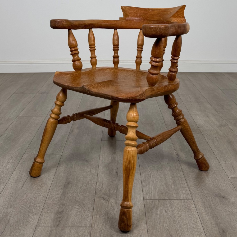 A solid oak smokers bow armchair featuring a shaped saddle seat, turned spindles, curved handrests, and splayed legs.