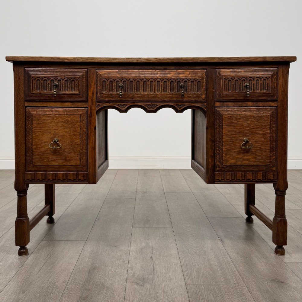 An Edwardian carved oak writing desk featuring five drawers with carved blind arcading, turned legs, stretcher supports, and brass handles.