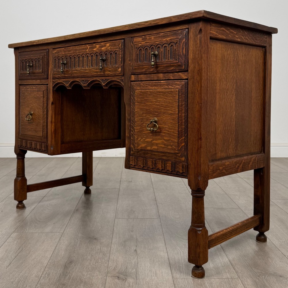 An Edwardian carved oak writing desk featuring five drawers with carved blind arcading, turned legs, stretcher supports, and brass handles. View II
