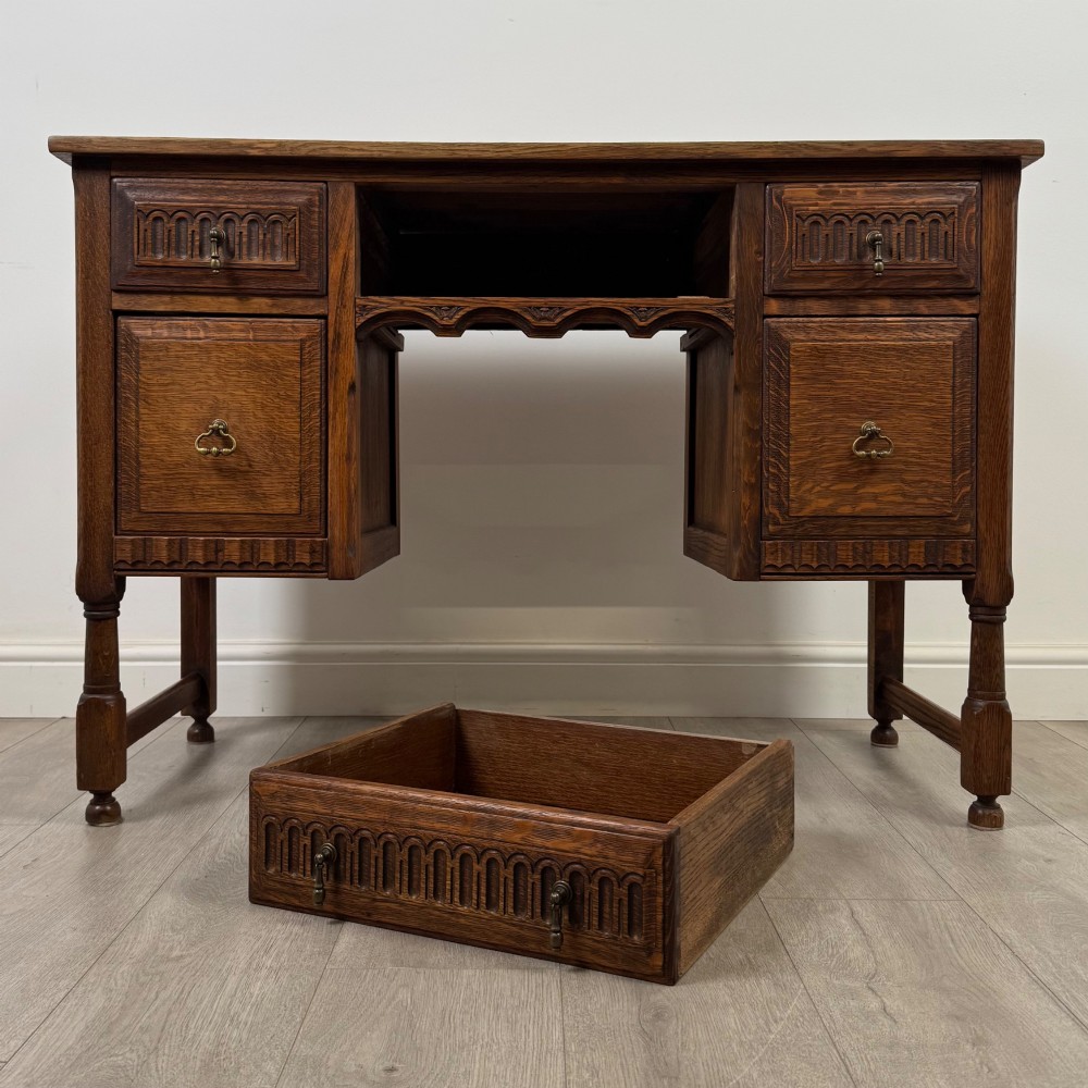 An Edwardian carved oak writing desk featuring five drawers with carved blind arcading, turned legs, stretcher supports, and brass handles. View X