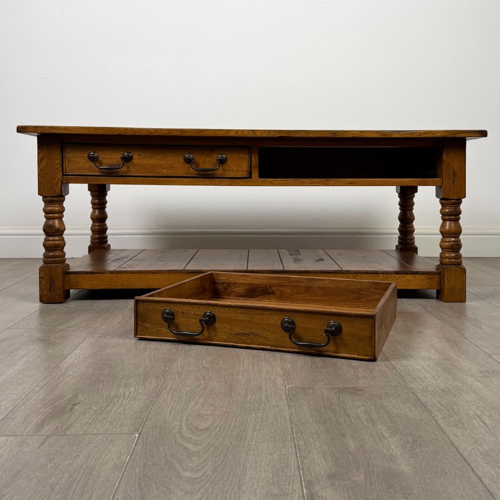 A low rectangular oak coffee table featuring two drawers with metal drop handles, turned legs, and an open lower plank shelf. View X