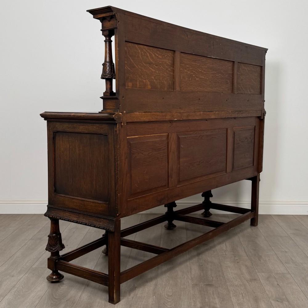 A large 19th-century dark carved oak sideboard featuring a raised upper gallery with carved panels, geometric cupboard doors, central drawers, and turned legs joined by stretchers. View V