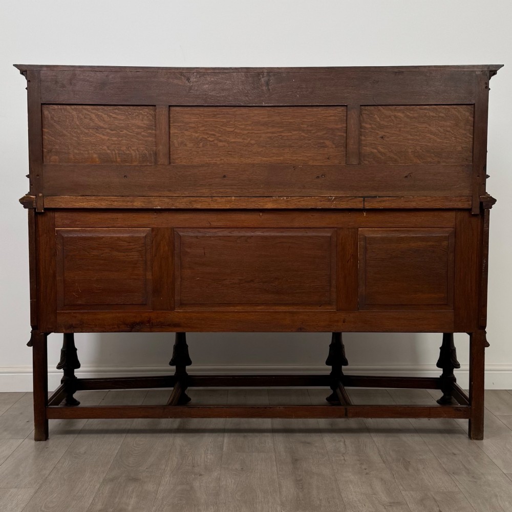 A large 19th-century dark carved oak sideboard featuring a raised upper gallery with carved panels, geometric cupboard doors, central drawers, and turned legs joined by stretchers. View VI