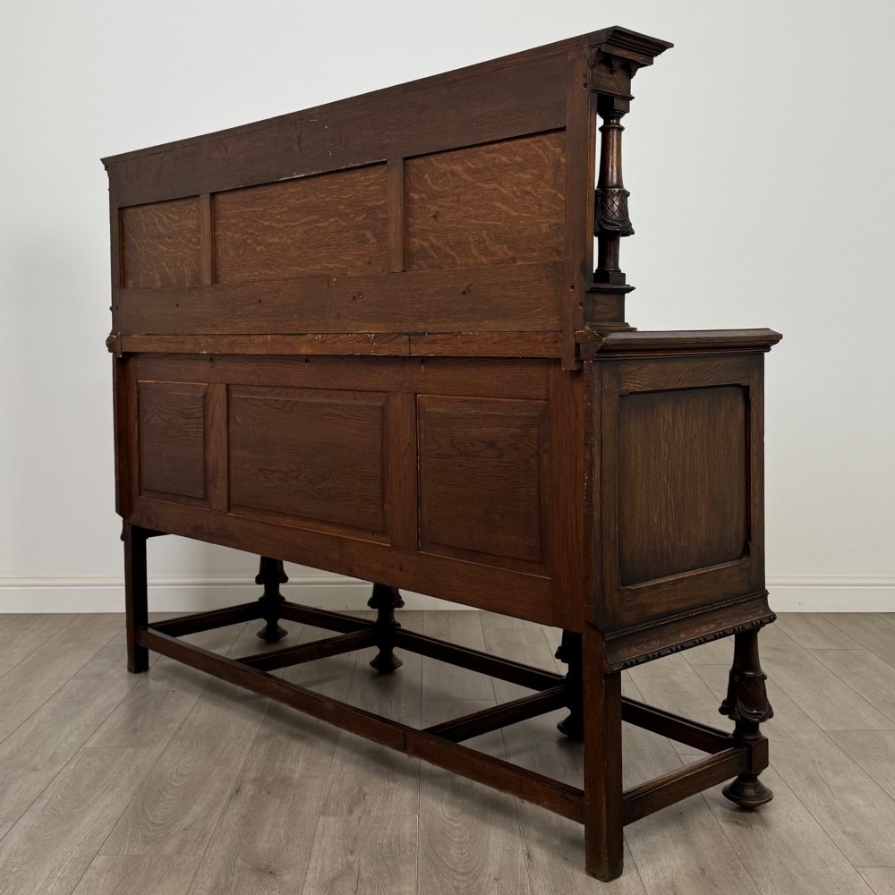 A large 19th-century dark carved oak sideboard featuring a raised upper gallery with carved panels, geometric cupboard doors, central drawers, and turned legs joined by stretchers. View VII