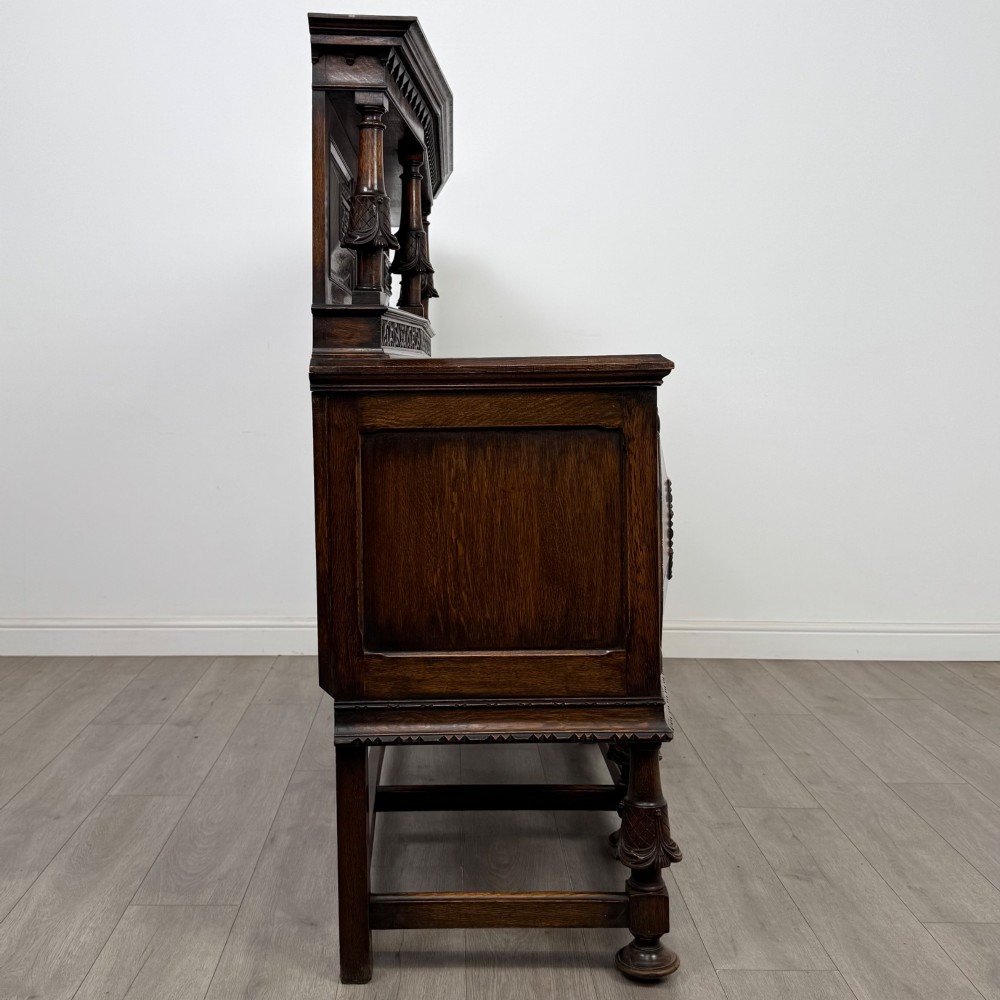 A large 19th-century dark carved oak sideboard featuring a raised upper gallery with carved panels, geometric cupboard doors, central drawers, and turned legs joined by stretchers. View VIII