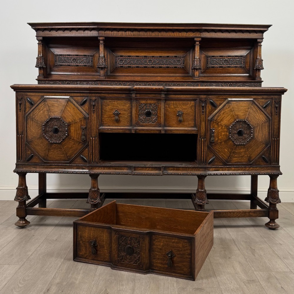 A large 19th-century dark carved oak sideboard featuring a raised upper gallery with carved panels, geometric cupboard doors, central drawers, and turned legs joined by stretchers. View XI