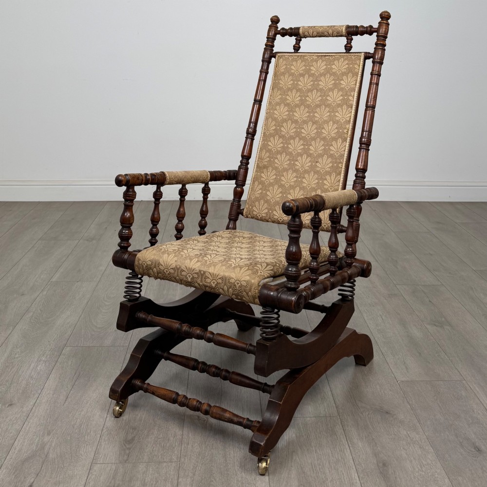 An American 20th-century platform rocking chair crafted from dark-stained beech with turned spindle arms, patterned gold upholstery, and a spring-loaded base on brass castors.