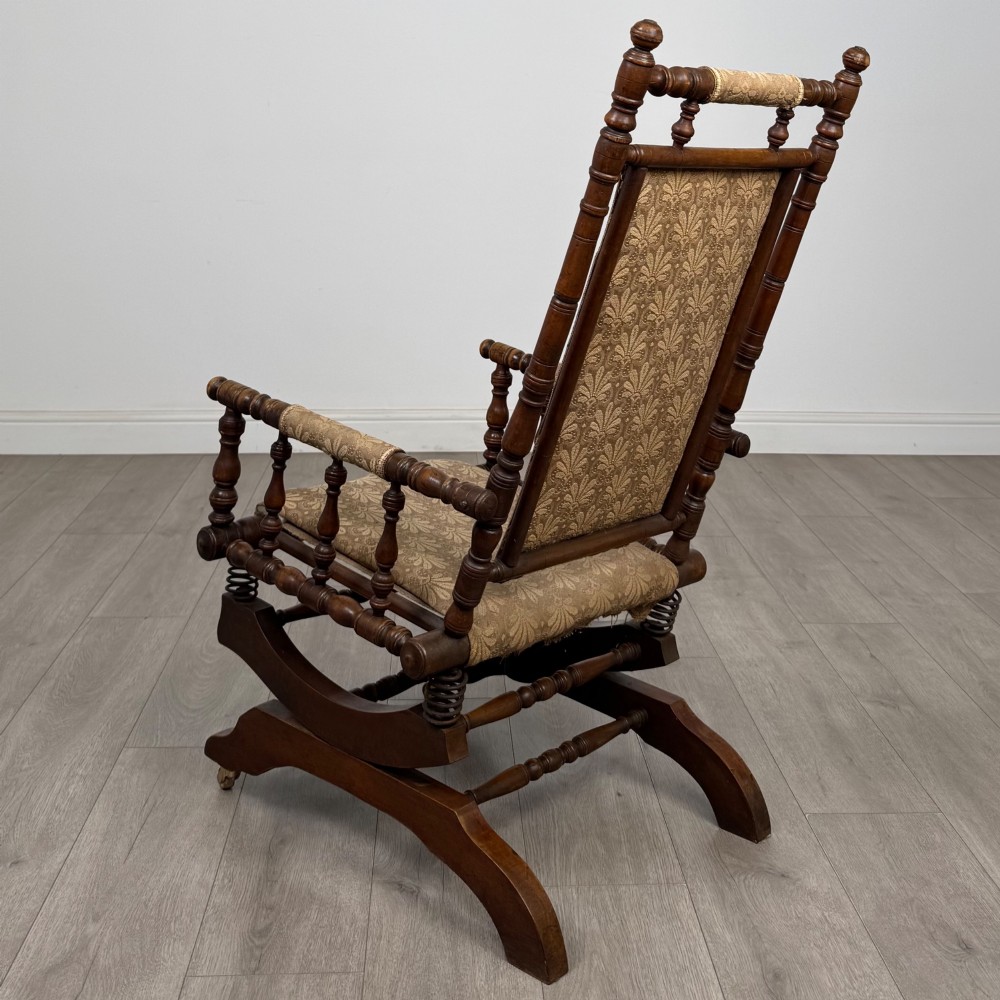 An American 20th-century platform rocking chair crafted from dark-stained beech with turned spindle arms, patterned gold upholstery, and a spring-loaded base on brass castors. View III