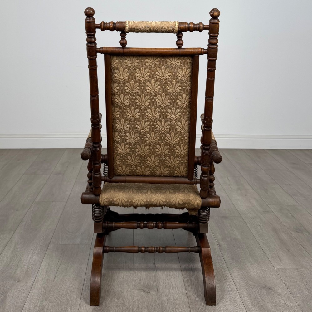 An American 20th-century platform rocking chair crafted from dark-stained beech with turned spindle arms, patterned gold upholstery, and a spring-loaded base on brass castors. View IV