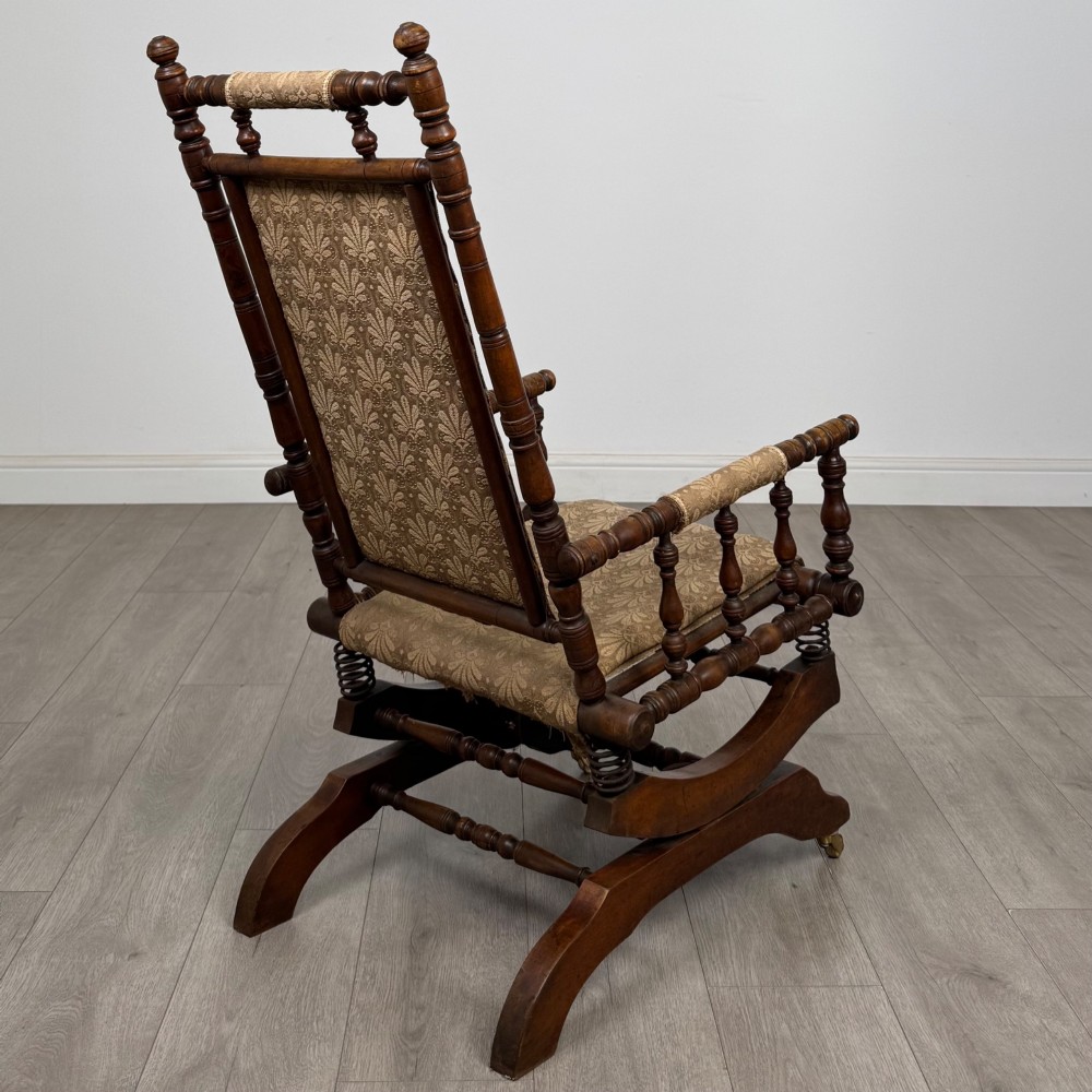 An American 20th-century platform rocking chair crafted from dark-stained beech with turned spindle arms, patterned gold upholstery, and a spring-loaded base on brass castors. View V