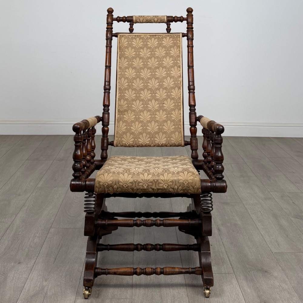An American 20th-century platform rocking chair crafted from dark-stained beech with turned spindle arms, patterned gold upholstery, and a spring-loaded base on brass castors. View VIII