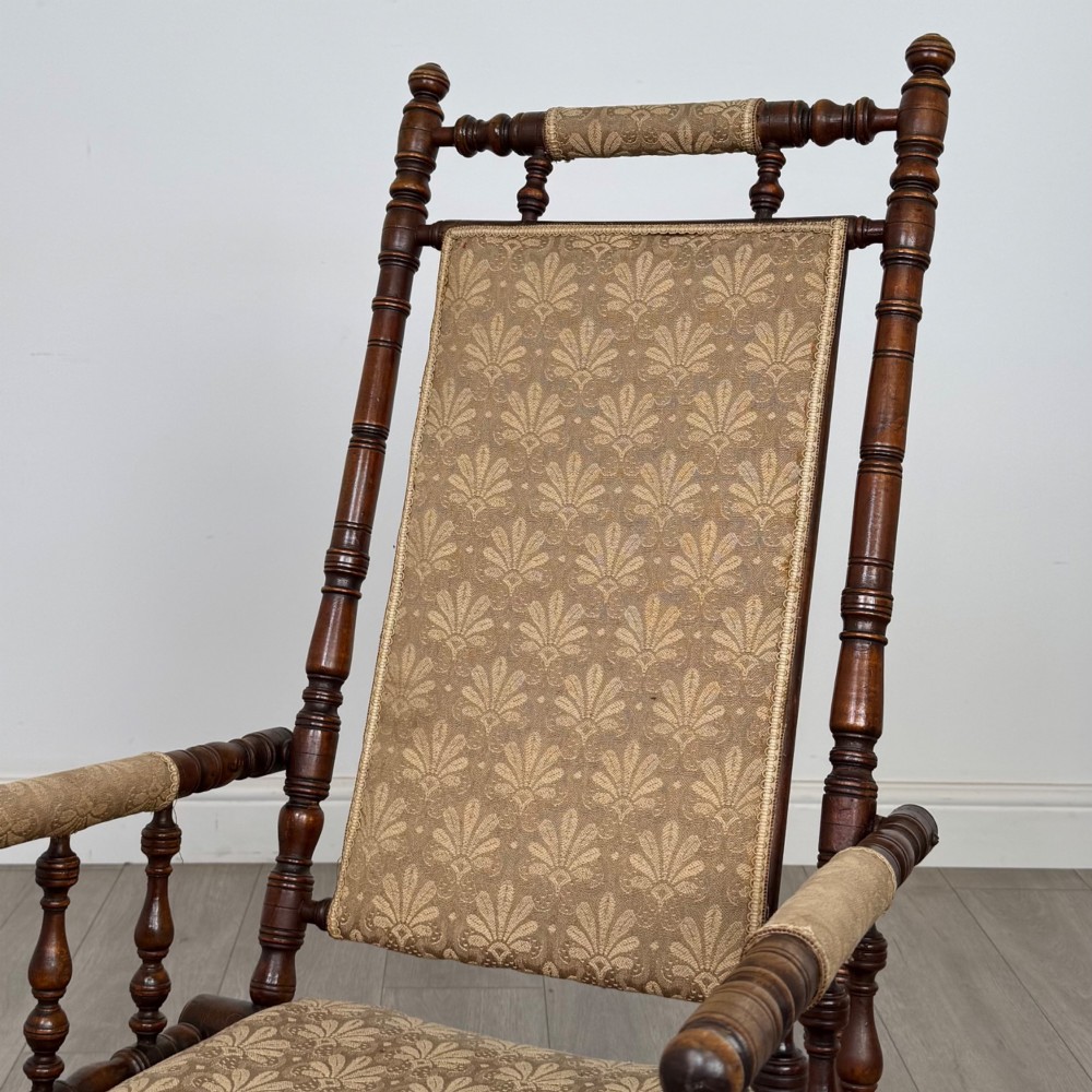 An American 20th-century platform rocking chair crafted from dark-stained beech with turned spindle arms, patterned gold upholstery, and a spring-loaded base on brass castors. View IX