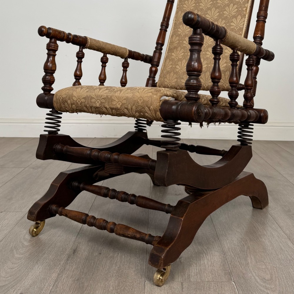 An American 20th-century platform rocking chair crafted from dark-stained beech with turned spindle arms, patterned gold upholstery, and a spring-loaded base on brass castors. View X