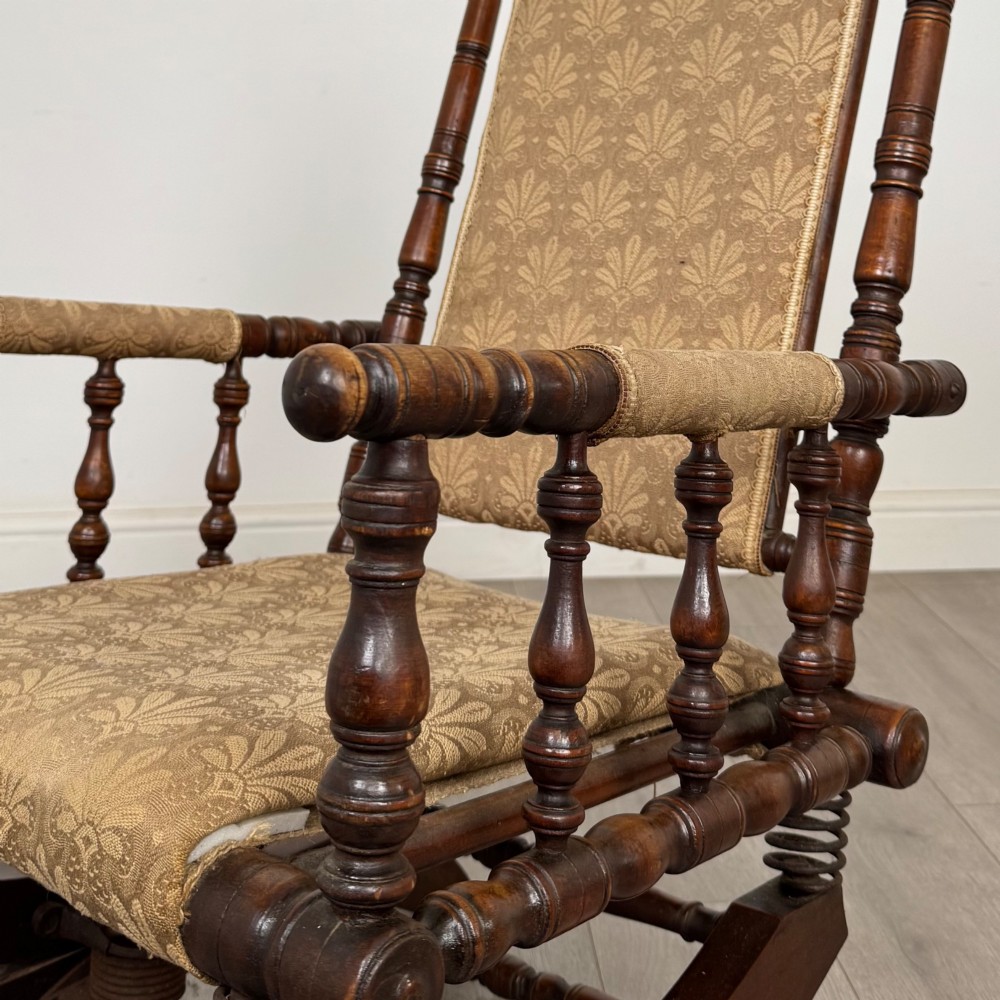 An American 20th-century platform rocking chair crafted from dark-stained beech with turned spindle arms, patterned gold upholstery, and a spring-loaded base on brass castors. View XI