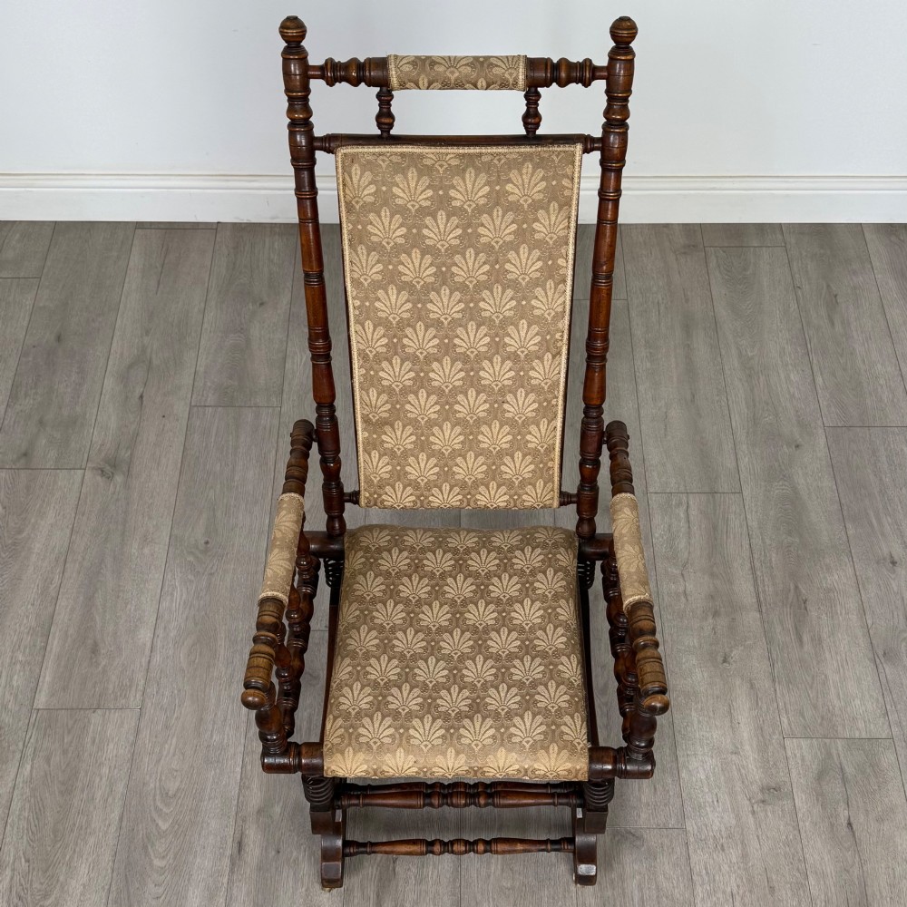 An American 20th-century platform rocking chair crafted from dark-stained beech with turned spindle arms, patterned gold upholstery, and a spring-loaded base on brass castors. View XII