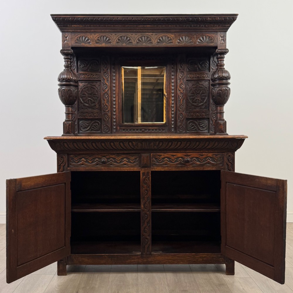 A 19th-century dark carved oak Victorian sideboard featuring an upper mirrored shelf section supported by turned bulbous pilasters above two carved drawers and cupboard doors with rosette reliefs. View II
