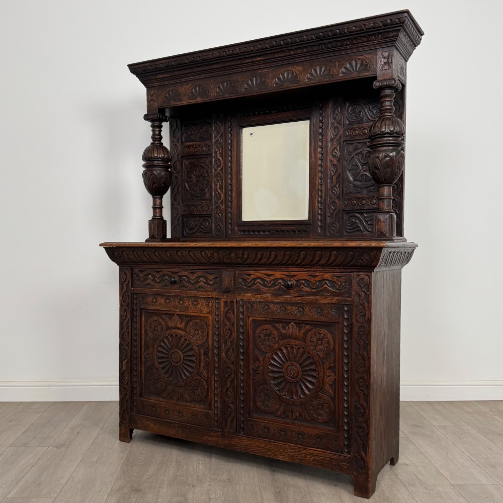 A 19th-century dark carved oak Victorian sideboard featuring an upper mirrored shelf section supported by turned bulbous pilasters above two carved drawers and cupboard doors with rosette reliefs. View III