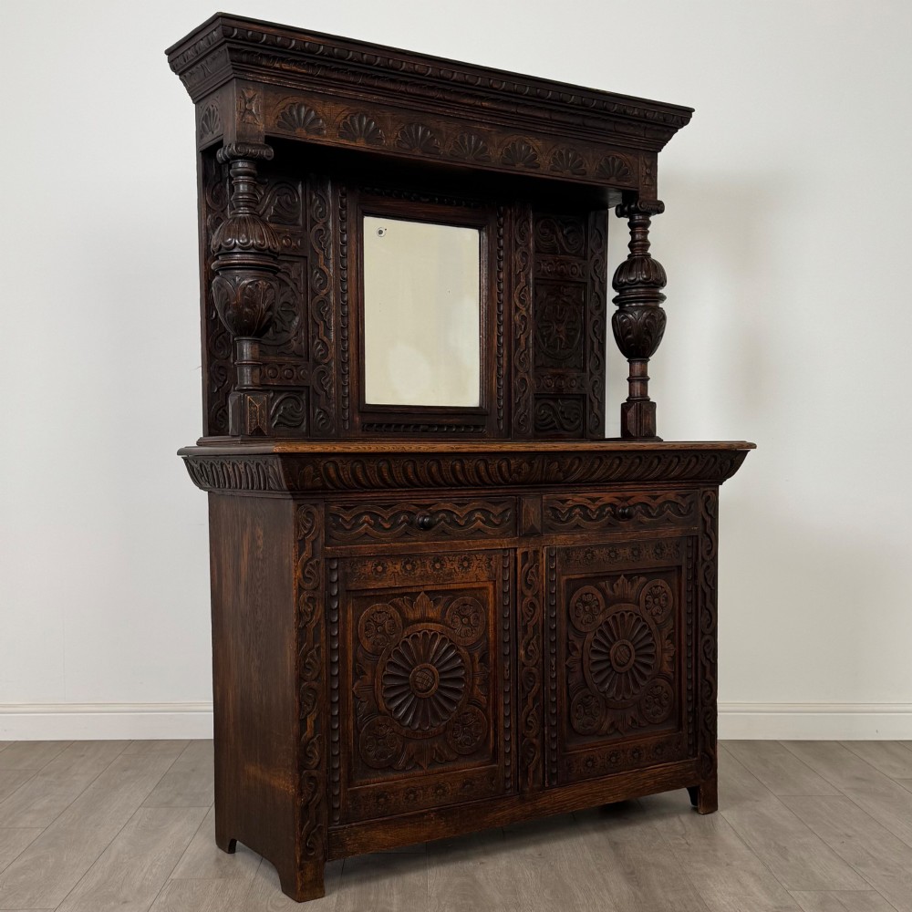 A 19th-century dark carved oak Victorian sideboard featuring an upper mirrored shelf section supported by turned bulbous pilasters above two carved drawers and cupboard doors with rosette reliefs. View IX