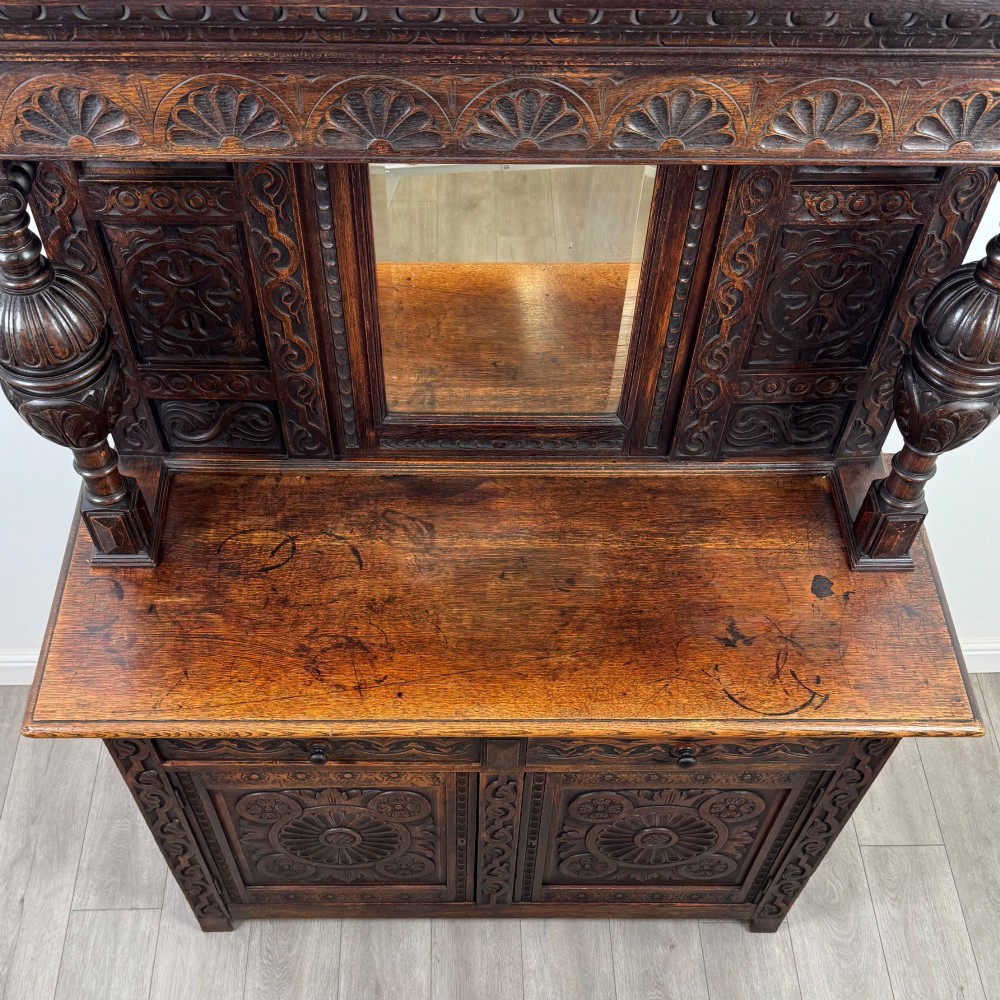 A 19th-century dark carved oak Victorian sideboard featuring an upper mirrored shelf section supported by turned bulbous pilasters above two carved drawers and cupboard doors with rosette reliefs. View X