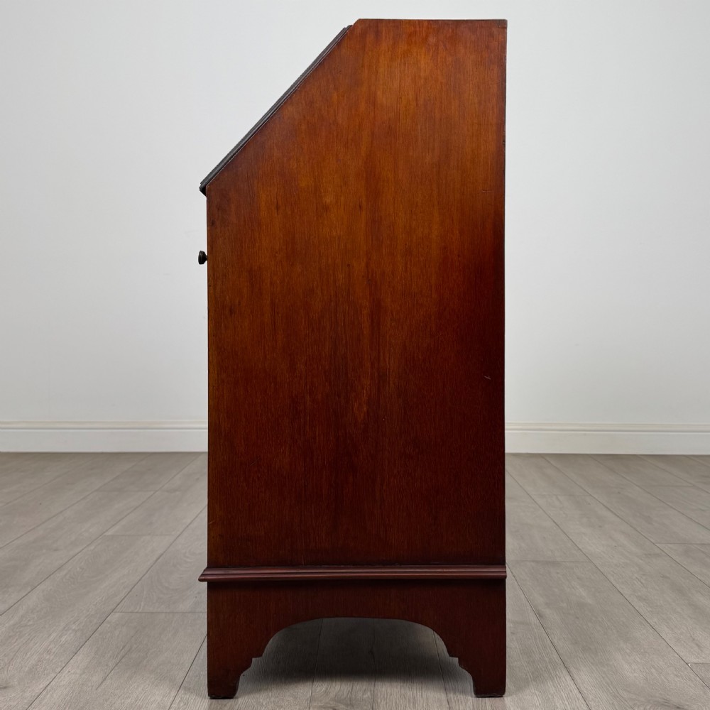 An Edwardian mahogany writing bureau featuring a slant front top, three long drawers with brass drop handles, and shaped bracket feet. View IV