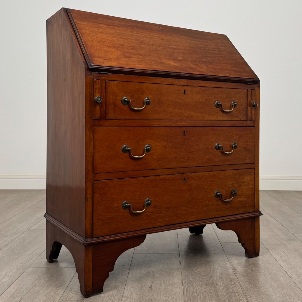 An Edwardian mahogany writing bureau featuring a slant front top, three long drawers with brass drop handles, and shaped bracket feet. View IX