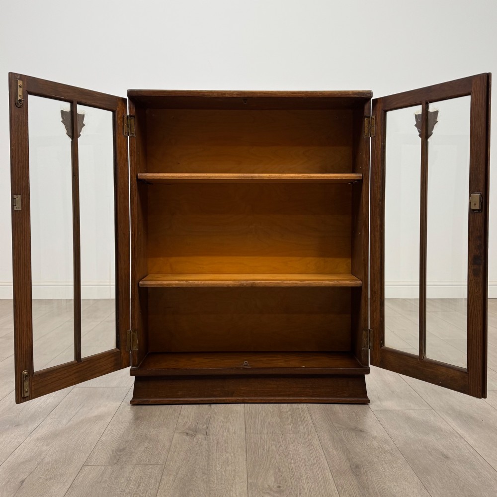 An Edwardian dark oak display cabinet with two glazed doors featuring vertical glazing bars topped with carved floral details. View II