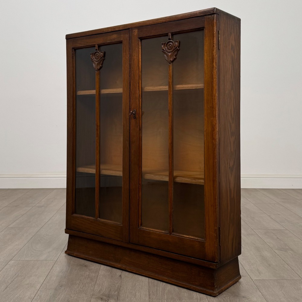 An Edwardian dark oak display cabinet with two glazed doors featuring vertical glazing bars topped with carved floral details. View III