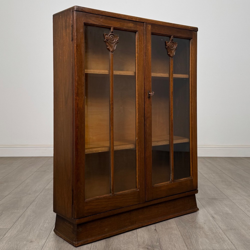 An Edwardian dark oak display cabinet with two glazed doors featuring vertical glazing bars topped with carved floral details. View IX