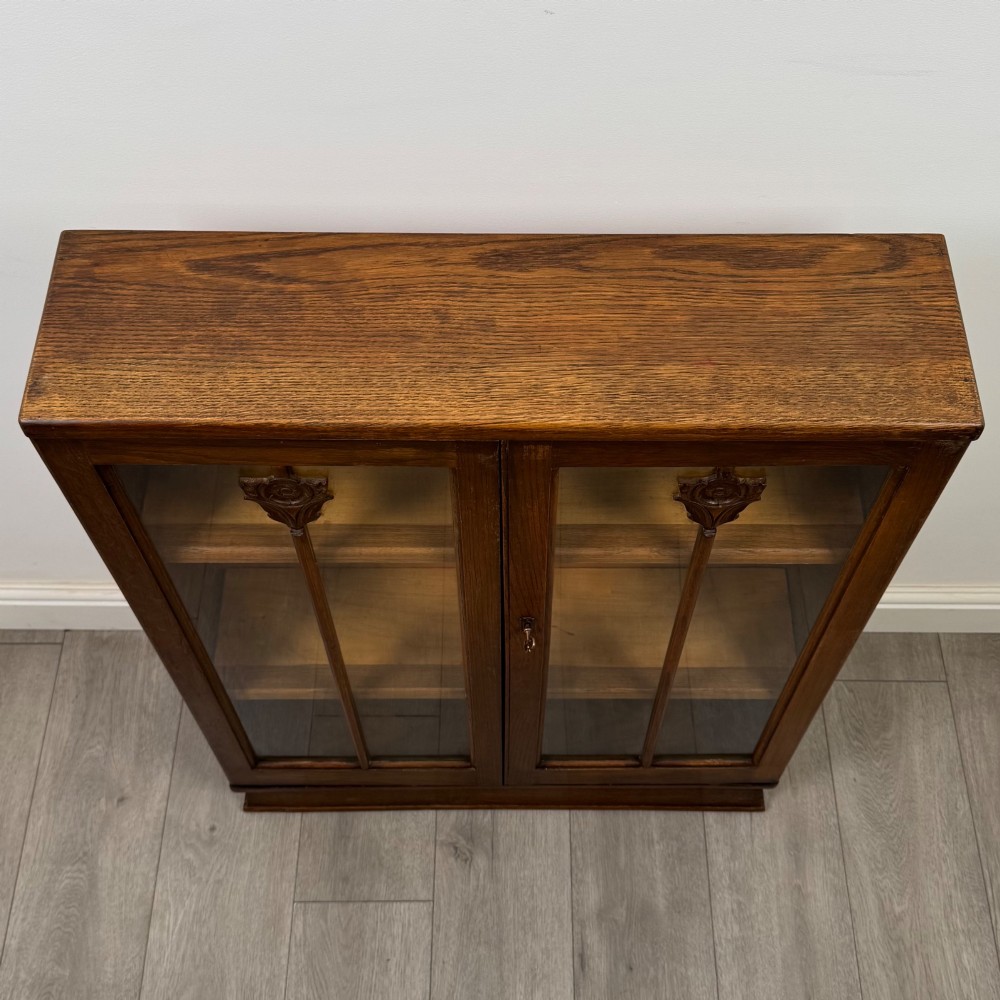 An Edwardian dark oak display cabinet with two glazed doors featuring vertical glazing bars topped with carved floral details. View X