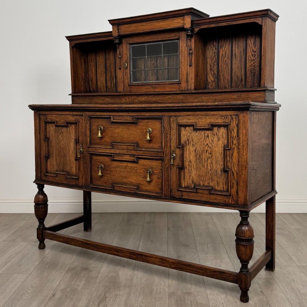 A 20th-century Jacobean style dark oak sideboard with a central leaded glass cupboard, geometric moulded drawers and doors, turned legs, and stretchers. View III