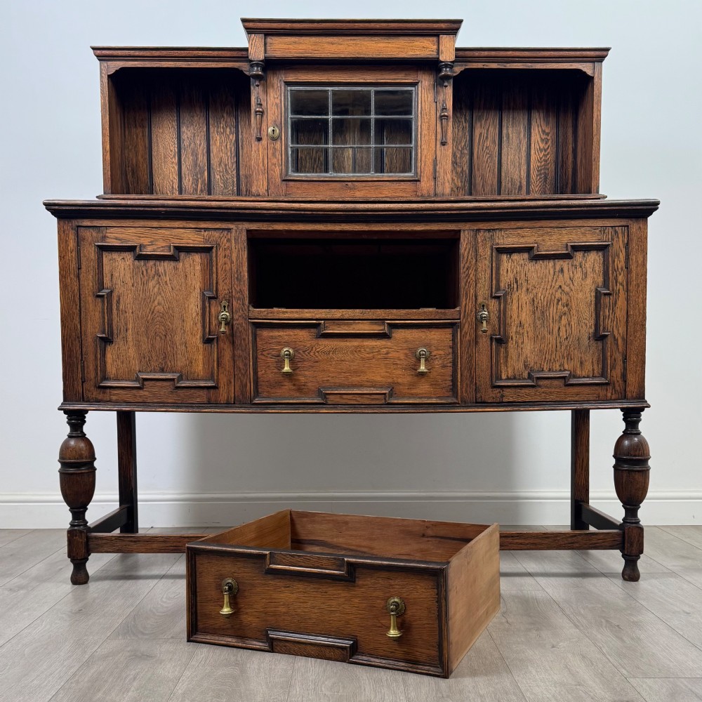 A 20th-century Jacobean style dark oak sideboard with a central leaded glass cupboard, geometric moulded drawers and doors, turned legs, and stretchers. View XI