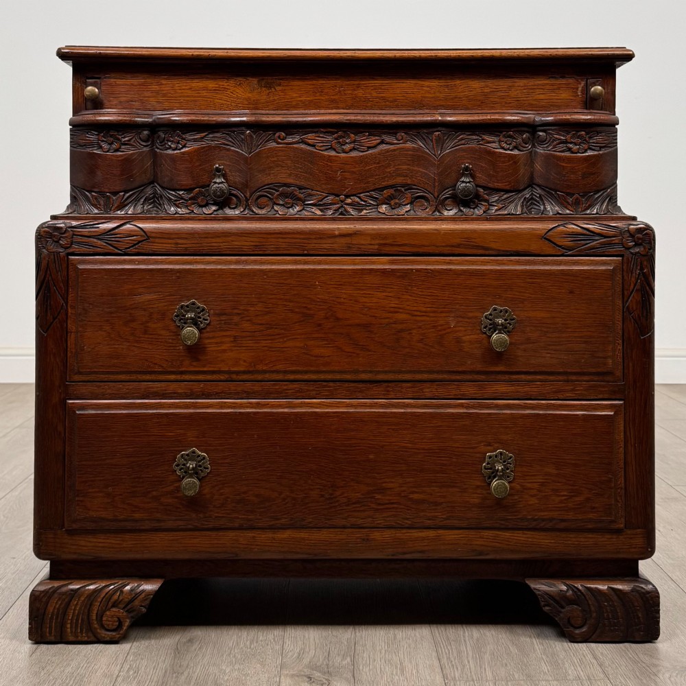 A dark carved oak chest of drawers featuring three drawers, floral relief carvings, and brass pendant handles set on carved bracket feet.