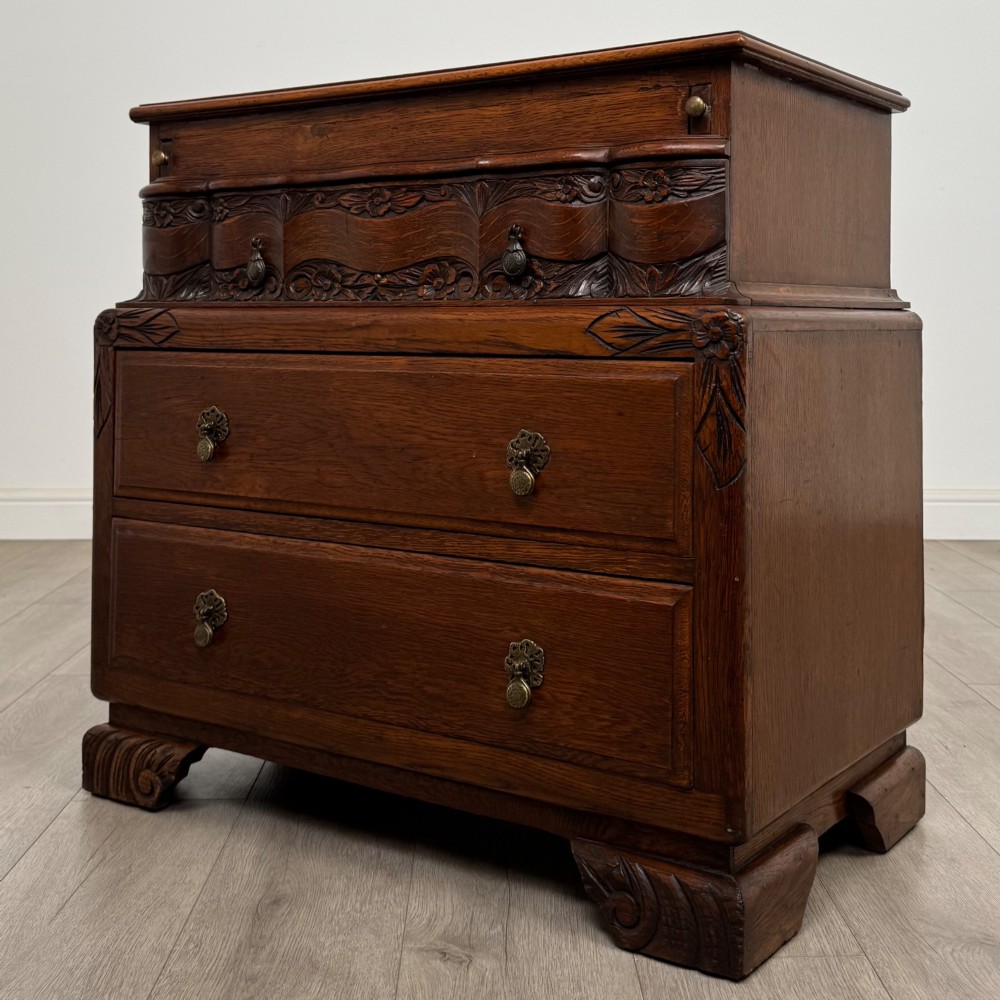A dark carved oak chest of drawers featuring three drawers, floral relief carvings, and brass pendant handles set on carved bracket feet. View II