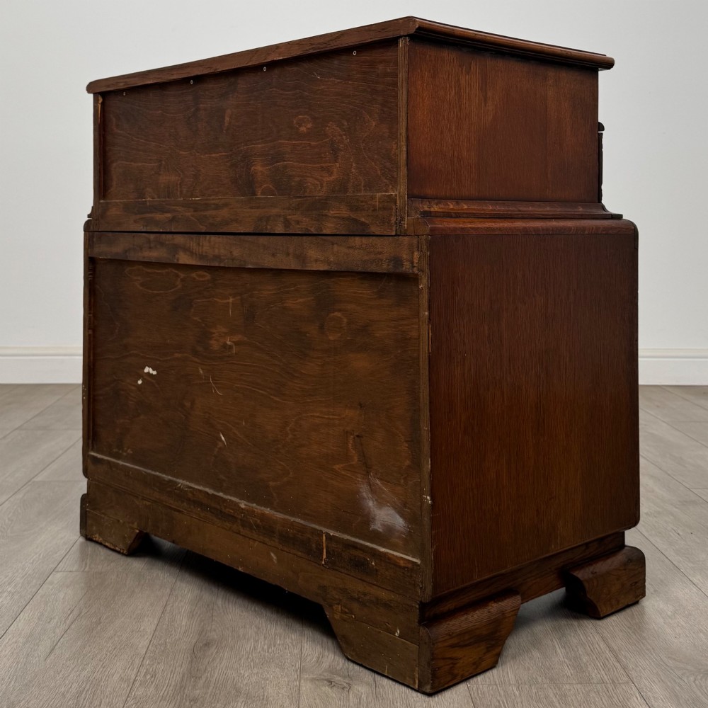 A dark carved oak chest of drawers featuring three drawers, floral relief carvings, and brass pendant handles set on carved bracket feet. View VI