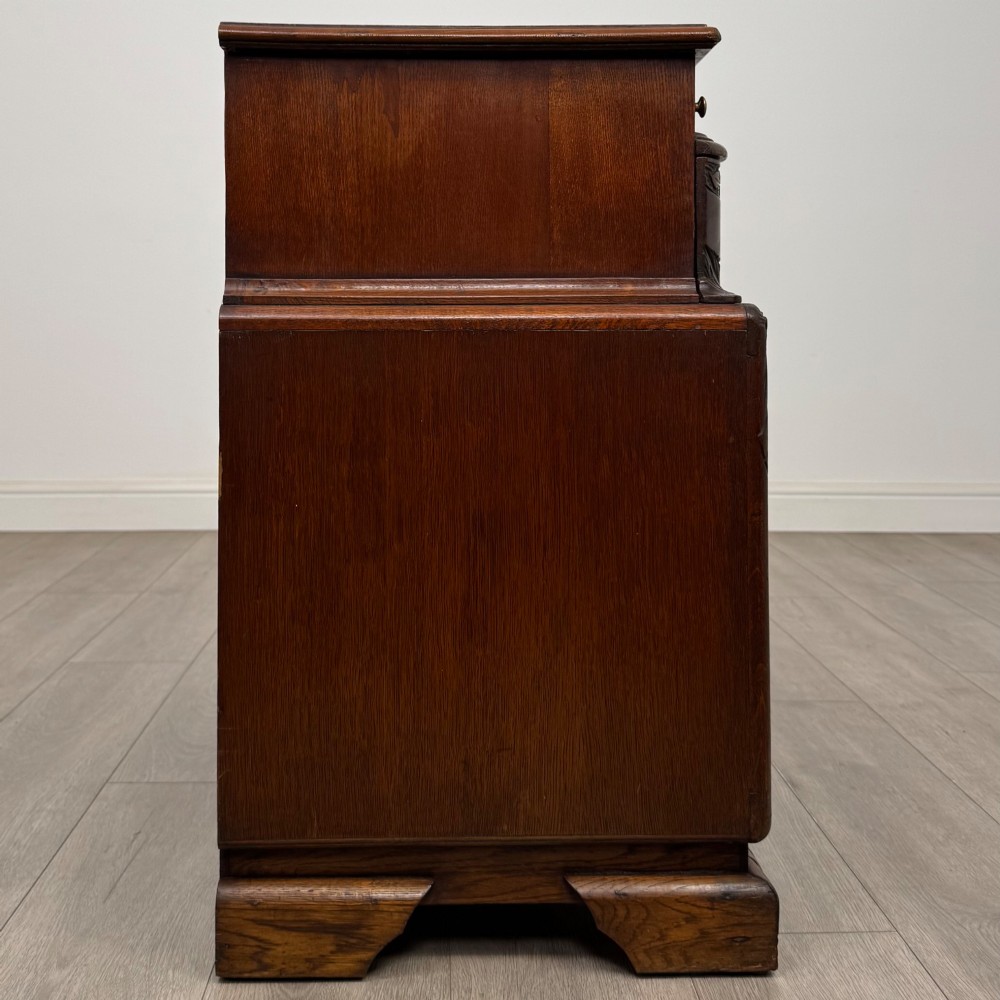 A dark carved oak chest of drawers featuring three drawers, floral relief carvings, and brass pendant handles set on carved bracket feet. View VII