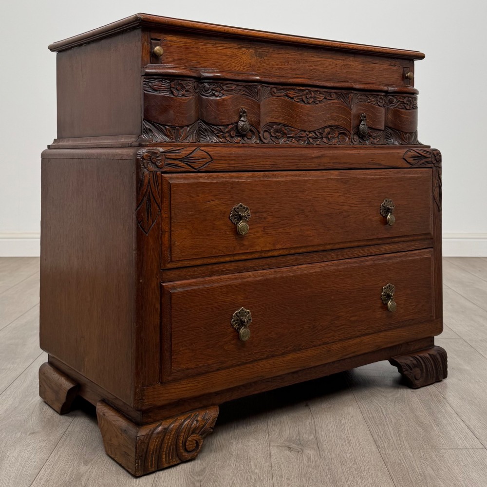 A dark carved oak chest of drawers featuring three drawers, floral relief carvings, and brass pendant handles set on carved bracket feet. View VIII