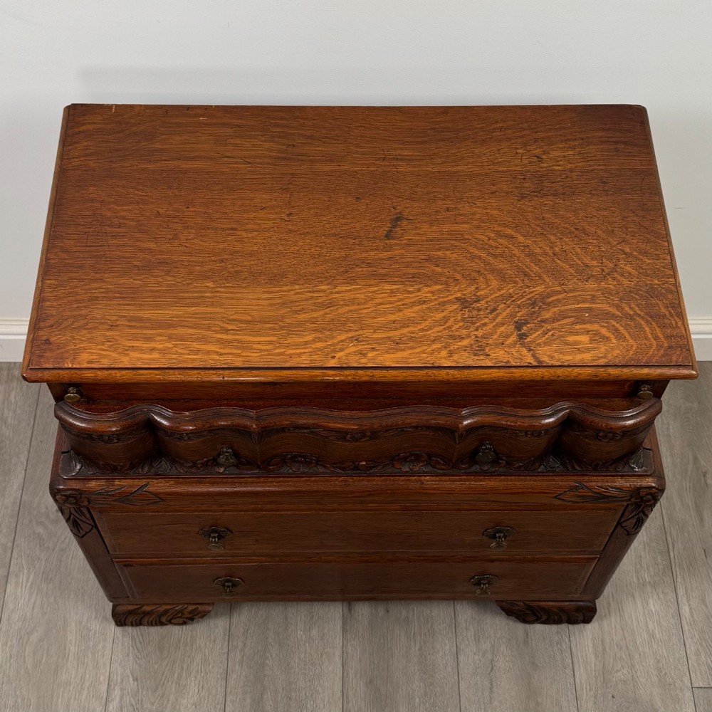 A dark carved oak chest of drawers featuring three drawers, floral relief carvings, and brass pendant handles set on carved bracket feet. View IX