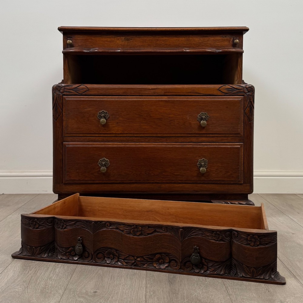 A dark carved oak chest of drawers featuring three drawers, floral relief carvings, and brass pendant handles set on carved bracket feet. View X